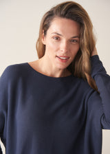A woman stands against a plain off-white wall wearing a navy blue knit top with a brown tie detail, white wide-leg trousers, and beige slip-on shoes.
