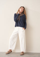 A woman stands against a plain off-white wall wearing a navy blue knit top with a brown tie detail, white wide-leg trousers, and beige slip-on shoes.