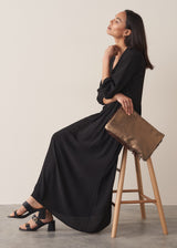 Woman sitting on a stool wearing black full length maxi dress with puffed 3/4 length sleeves, paired with gold clutch bag and black sandals