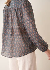 A woman wearing a dark blue long-sleeve blouse with a brown and rust-colored paisley print pattern and light trousers.