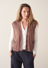 A woman wearing a sleeveless brown knit vest over a white long-sleeve top and dark blue jeans, standing against a plain light beige background.