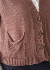 A woman wearing a sleeveless brown knit vest over a white long-sleeve top and dark blue jeans, standing against a plain light beige background.