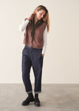A woman wearing a sleeveless brown knit vest over a white long-sleeve top and dark blue jeans, standing against a plain light beige background.