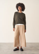 A woman wearing a khaki green crewneck sweater, layered over a white top and beige trousers.