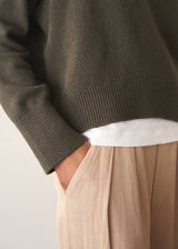 A woman wearing a khaki green crewneck sweater, layered over a white top and beige trousers.