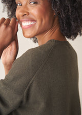 A woman wearing a khaki green crewneck sweater, layered over a white top and beige trousers.