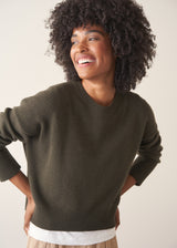 A woman wearing a khaki green crewneck sweater, layered over a white top and beige trousers.