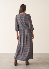 A woman wearing a dark blue midi dress with a brown and beige paisley pattern, featuring puff sleeves and a deep V-neckline, set against a plain beige background.