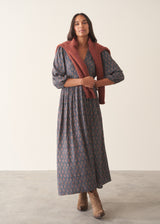 A woman wearing a dark blue midi dress with a brown and beige paisley pattern, featuring puff sleeves and a deep V-neckline, set against a plain beige background.