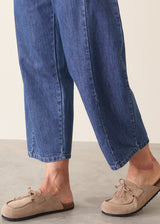 A person wearing blue denim cropped pants and taupe suede slip-on mule loafers, shown from the lower legs down against a neutral studio background.