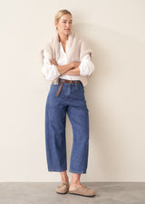 A woman stands against a plain beige wall wearing blue cropped wide-leg jeans, a white lace-trim blouse, a beige knit cardigan, and tan loafers.