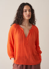 Woman wearing a bright orange cotton blouse and linen trousers against a plain background