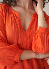 Woman wearing a bright orange cotton blouse and linen trousers against a plain background