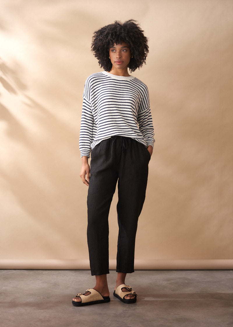 Woman wearing a light knit stripe crew neck sweater with black trousers, on a neutral background