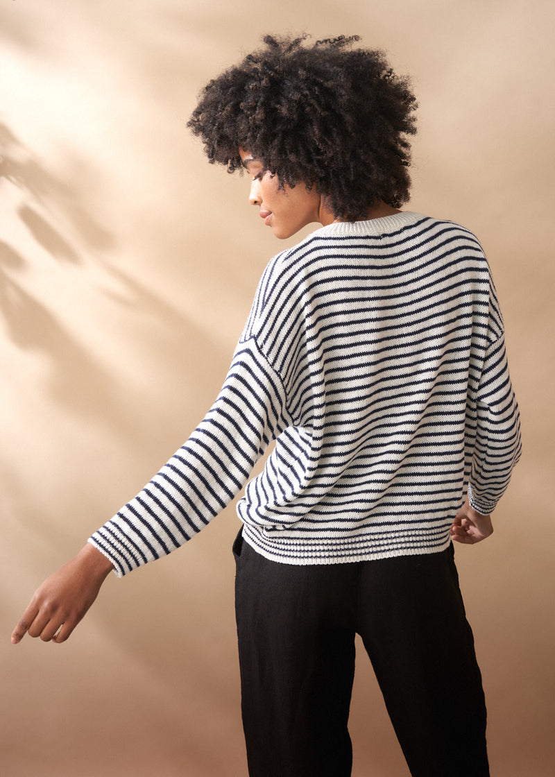 Woman wearing a light knit striped crew neck sweater with black trousers, facing a neutral backgroud