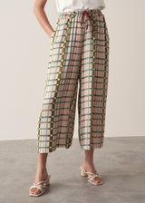 A woman models silk blend wide-leg trousers in a check print, paired with a sleeveless satin top and strappy sandals, standing in front of a beige background