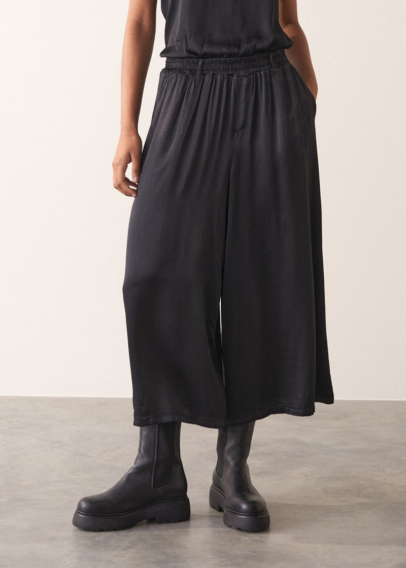 Woman wearing black satin wide leg cropped trousers against a plain background