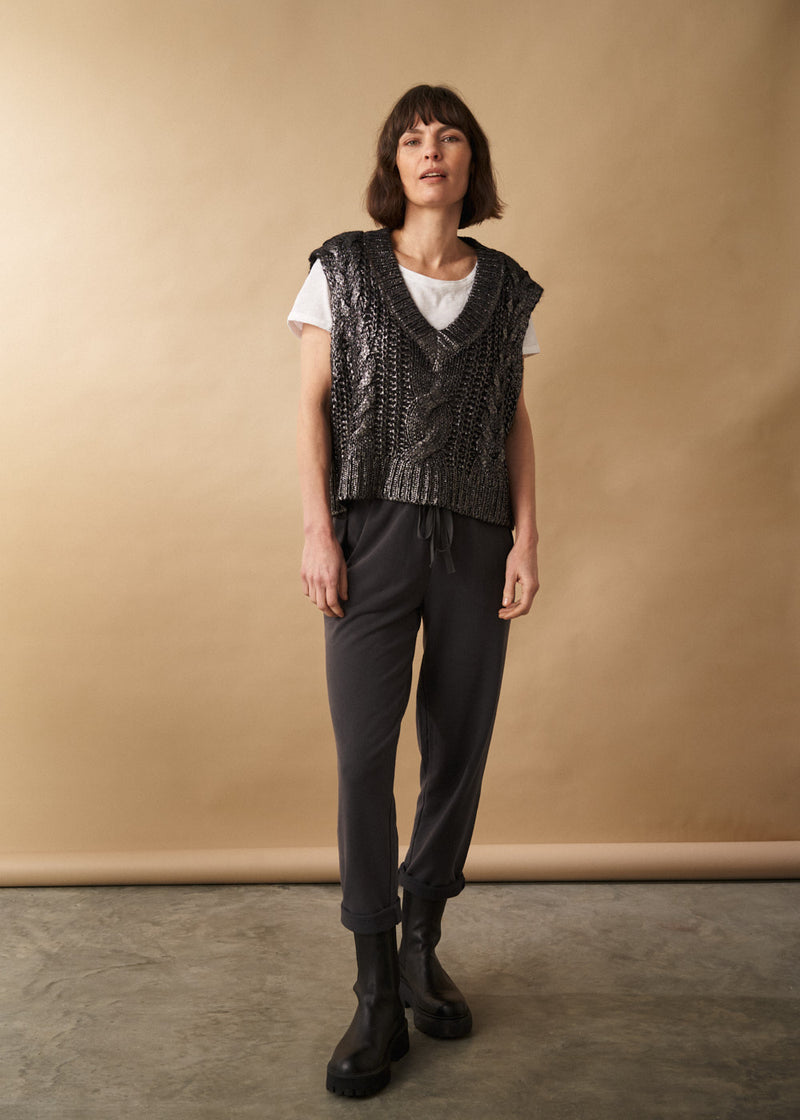 Woman wears a dark grey sleeveless jumper with white t-shirt and jersey jogging bottoms, with a neutral backdrop.