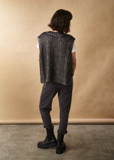 Woman wears a dark grey metallic silver cable knit vest, white t-shirt, jersey trousers and black leather boots facing a neutral background