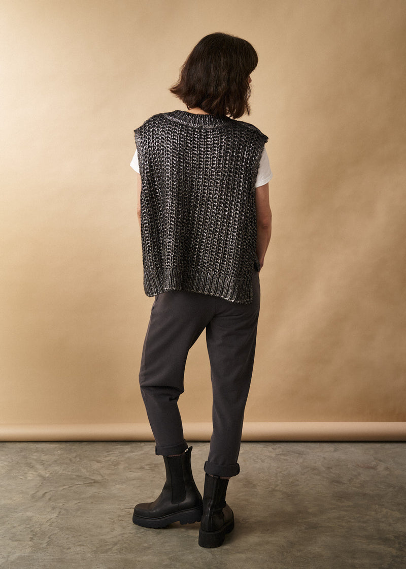 Woman wears a dark grey metallic silver cable knit vest, white t-shirt, jersey trousers and black leather boots facing a neutral background