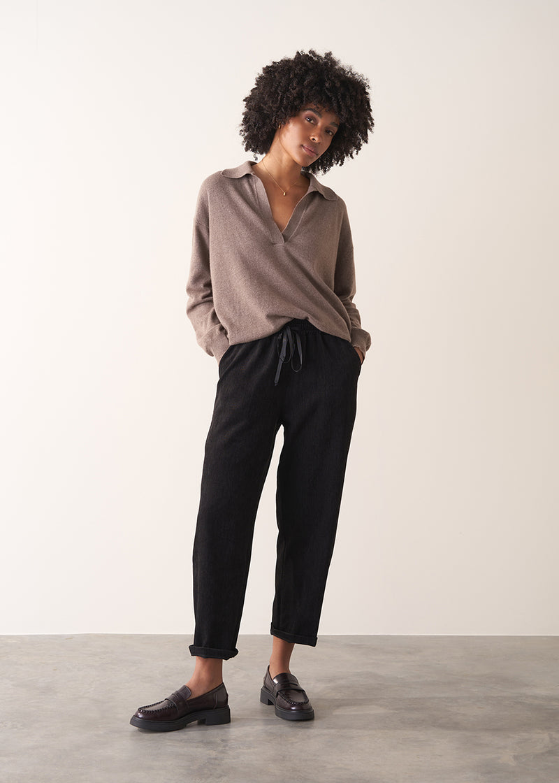 Woman wearing a light brown long-sleeve polo neck sweater and black drawstring trousers against a plain background