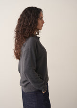 Woman wearing a dark grey polo neck collared jumper and denim wide leg jeans against a beige background