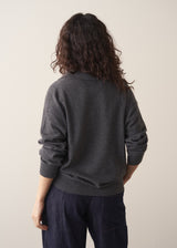 Woman wearing a dark grey polo neck collared jumper and denim wide leg jeans against a beige background
