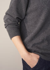 Woman wearing a dark grey polo neck collared jumper and denim wide leg jeans against a beige background