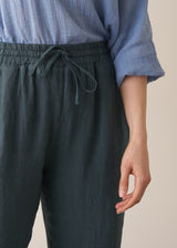 Close up of elasticated waist detail on a pair of dark green, tapered leg, ankle-length linen trousers, styled with a blue blouse