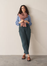 A woman leant against a wall, wearing a pair of dark green, tapered leg, ankle-length linen trousers with a blue blouse and brown sandals