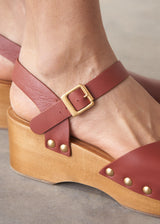 Close-up of a sandal with a wooden wedge sole and red strap with gold buckle.