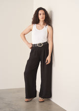 Woman wearing a white tank top and black wide-leg linen trousers against a plain background