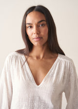 Woman wearing a white embroidered cotton blouse with a V-neck and black trousers against a plain background