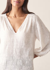 Woman wearing a white embroidered cotton blouse with a V-neck and black trousers against a plain background