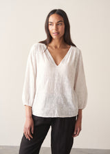Woman wearing a white embroidered cotton blouse with a V-neck and black trousers against a plain background
