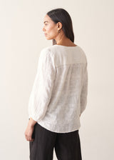 Woman wearing a white embroidered cotton blouse with a V-neck and black trousers against a plain background