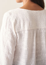 Woman wearing a white embroidered cotton blouse with a V-neck and black trousers against a plain background