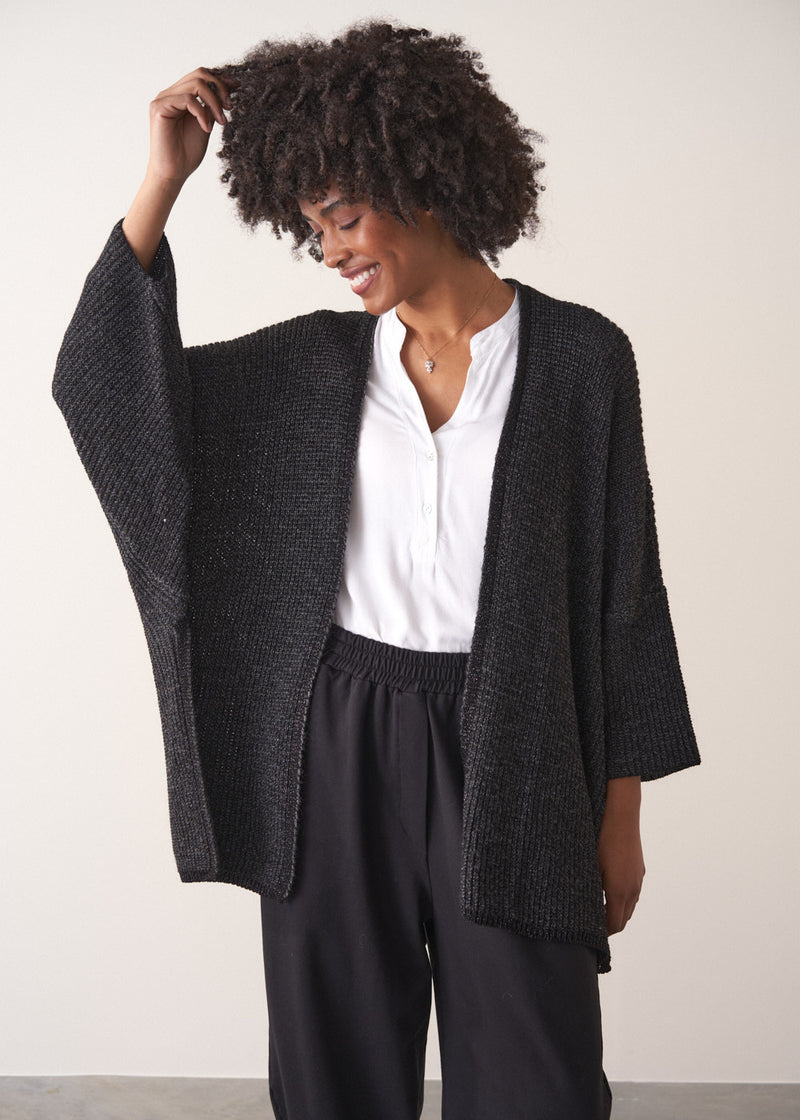 Woman wearing a dark gray cardigan over a white shirt and black trousers against a plain background