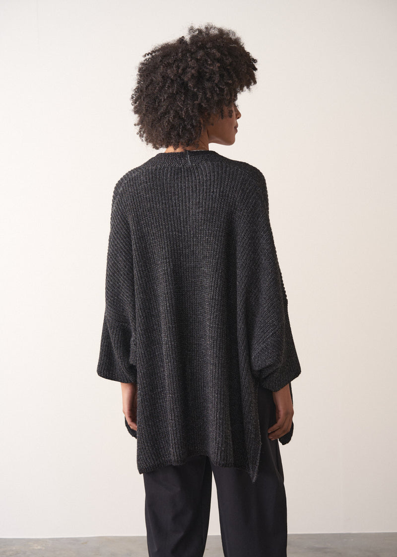 Woman wearing a dark gray cardigan over a white shirt and black trousers against a plain background