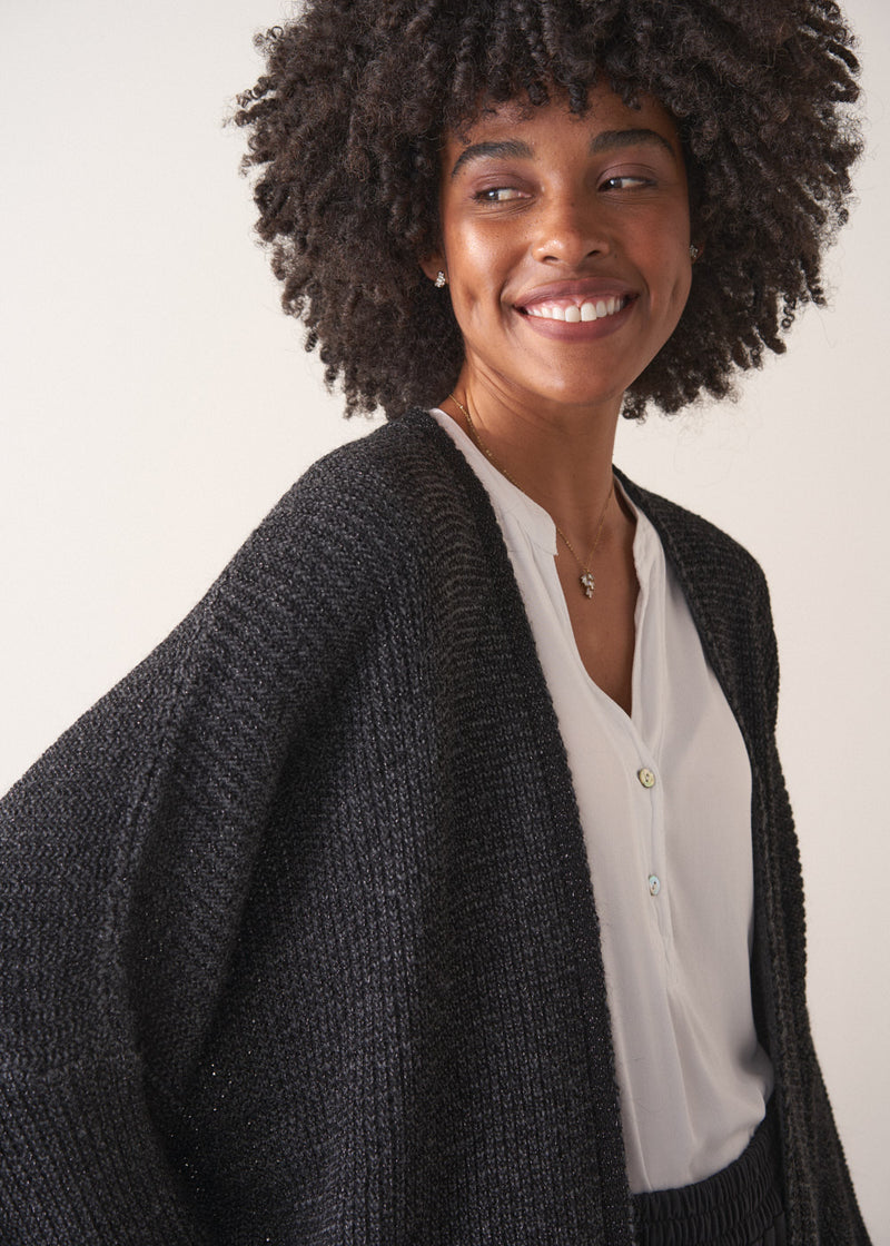 Woman wearing a dark gray cardigan over a white shirt and black trousers against a plain background