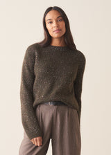 A woman wearing a dark khaki green sequin sweater with brown checked trousers and a black belt