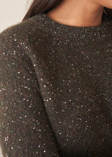 A woman wearing a dark khaki green sequin sweater with brown checked trousers and a black belt