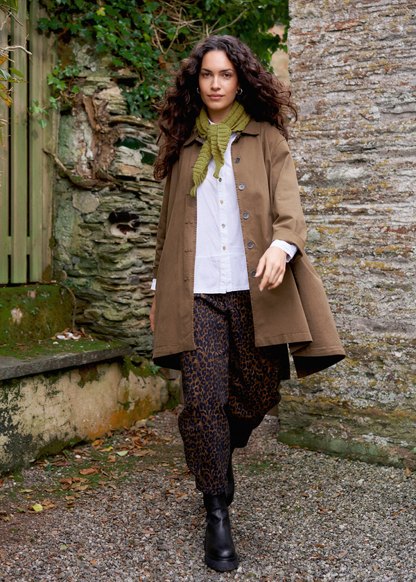 A woman walking through a garden, with a stone wall visible in the background. She wears a mid-length brown trench coat, a cotton blouse, leopard print trousers, and black Chelsea boots.