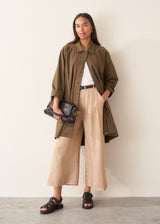 A woman wearing a mid-length khaki green utility trench coat over a white top and beige trousers.