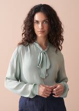 Woman wearing a light green blouse with a tie neckline against a beige background