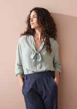 Woman wearing a light green blouse with a tie detail and dark blue jeans against a beige wall.