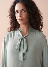 Woman wearing a light green blouse with a tie neckline against a beige background