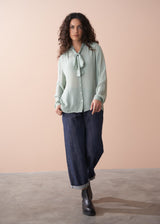 Woman wearing a light green blouse with a tie and dark blue pants against a beige background