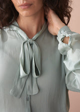 Woman wearing a light green blouse with a tie detail and ruffled sleeves.
