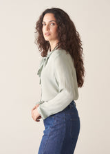 Woman wearing a light green blouse with a bow and blue jeans against a plain background